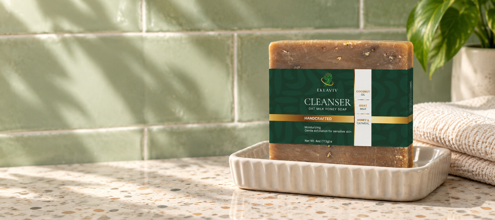 Dry wrapped Oat Milk and Honey Cleanser bar on a draining dish beside cotton towels and sage bathroom tile